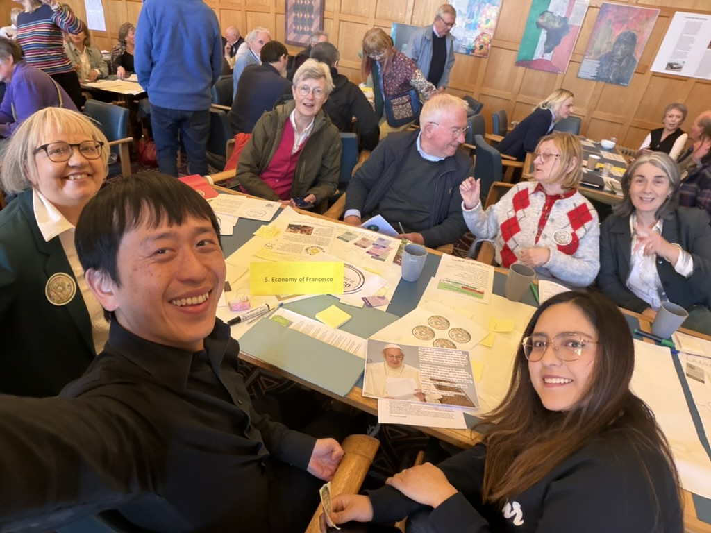 The photo features a group of participants gathered around the "Economy of Francesco" World Café table. It shows the specific tools from the workshop in action, including the sticky notes and large feedback sheets used to map out ideas. Additionally, a picture of Pope Francis is visible on the table, grounding the discussion in the movement's origins and his provocation that "this economy kills". It illustrates the "space for encounter" and the collaborative brainstorming that defined the session.